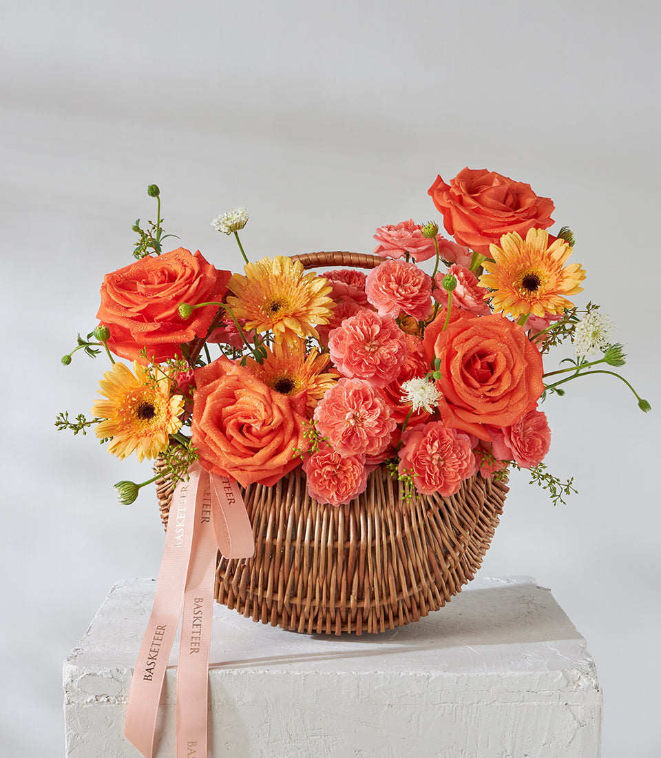 Vibrant orange roses, carnations and gerbera daisies in woven wicker basket with pink Basketeer ribbon