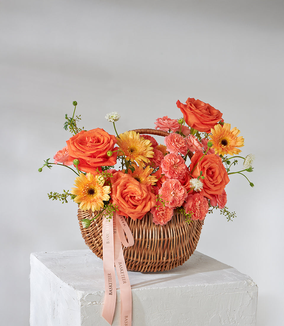 Vibrant orange roses, carnations and gerbera in woven basket with pink ribbons