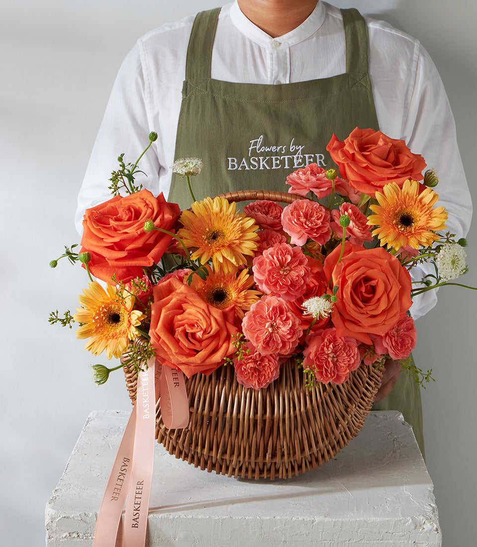 Vibrant orange roses, carnations, and gerbera daisies arranged in a wicker basket with green foliage and ribbon