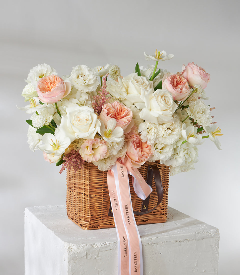 Peach and white garden roses in wicker basket with delicate filler blooms, light pink ribbon detail