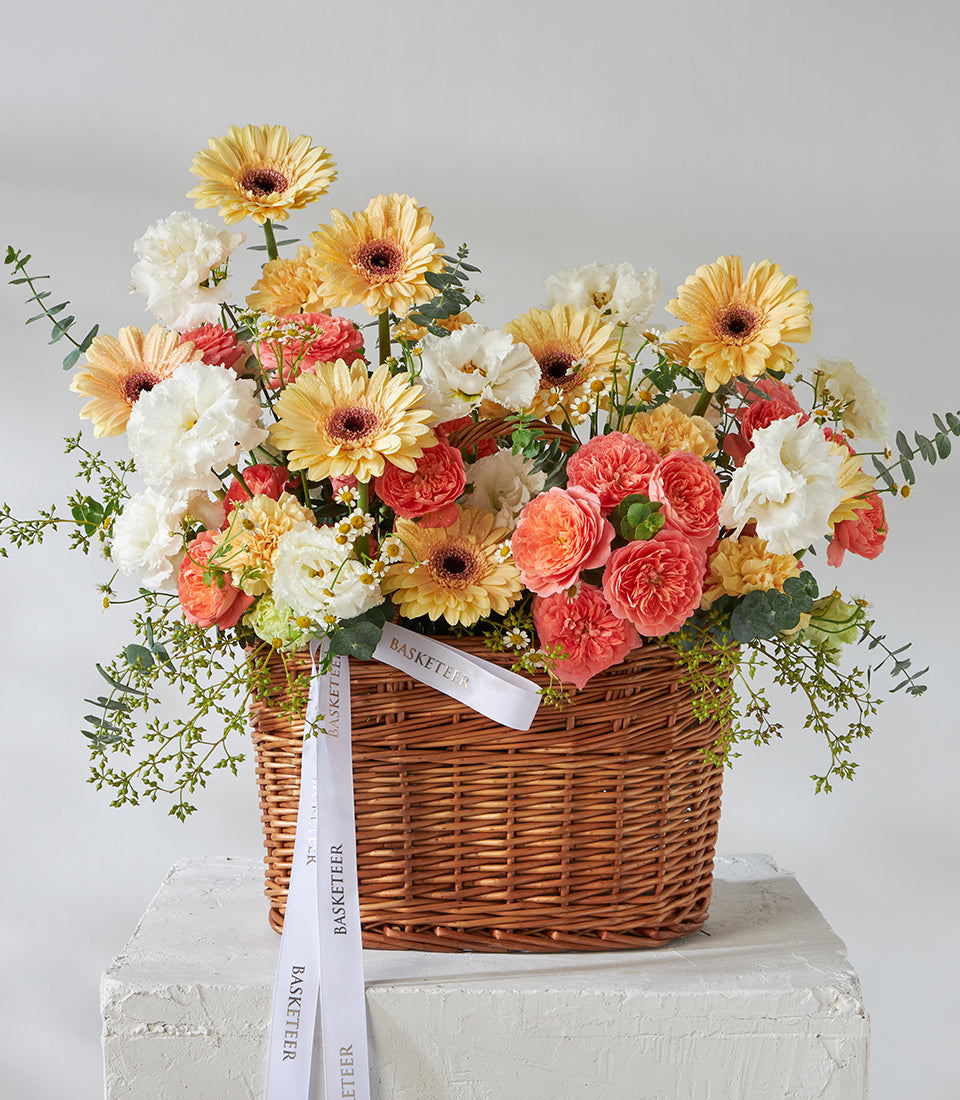 Peach roses, yellow gerberas, and white carnations in wicker basket with green foliage, birthday flowers
