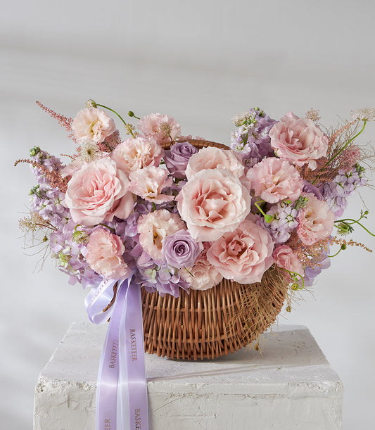 Blush pink roses and purple lisianthus in wicker basket with satin ribbons