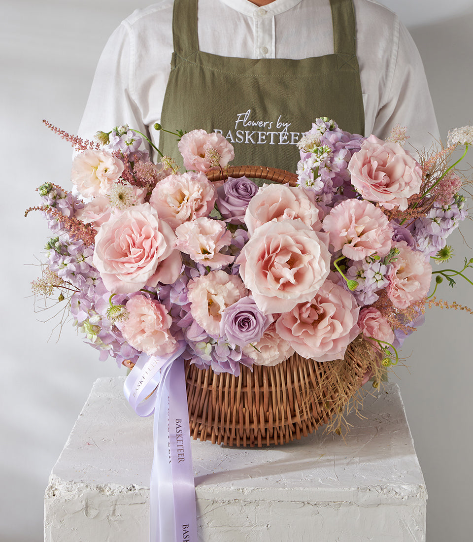 Blush pink roses and purple lisianthus in wicker basket with green foliage and ribbon bow