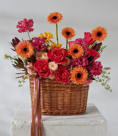 Red roses and orange gerbera daisies in woven wicker basket with pink carnations and foliage