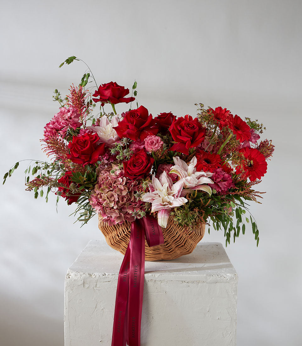 Red roses and pink lilies in woven basket with hydrangeas and greenery, luxury gift arrangement