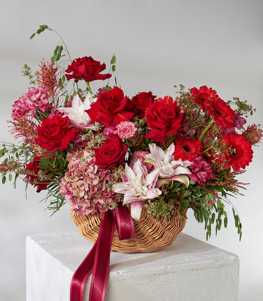 Red roses and pink lilies in a woven basket with hydrangea and filler flowers, accented with satin ribbon