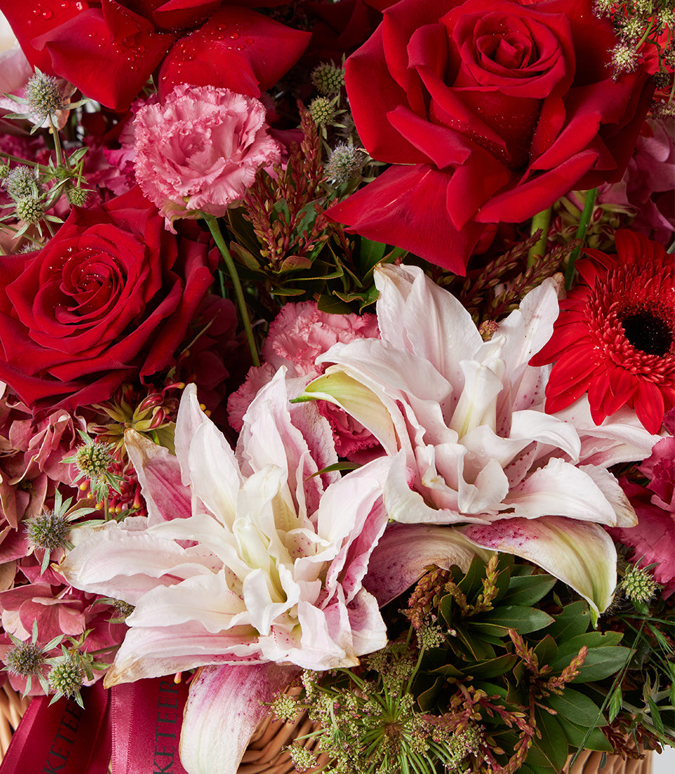 Deep red roses and pale pink lilies in a woven basket with carnations and hydrangeas, accented with green foliage