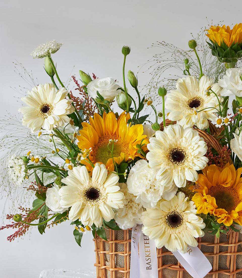 Bright yellow sunflowers and white gerbera daisies in a woven basket with green foliage, birthday flower gift
