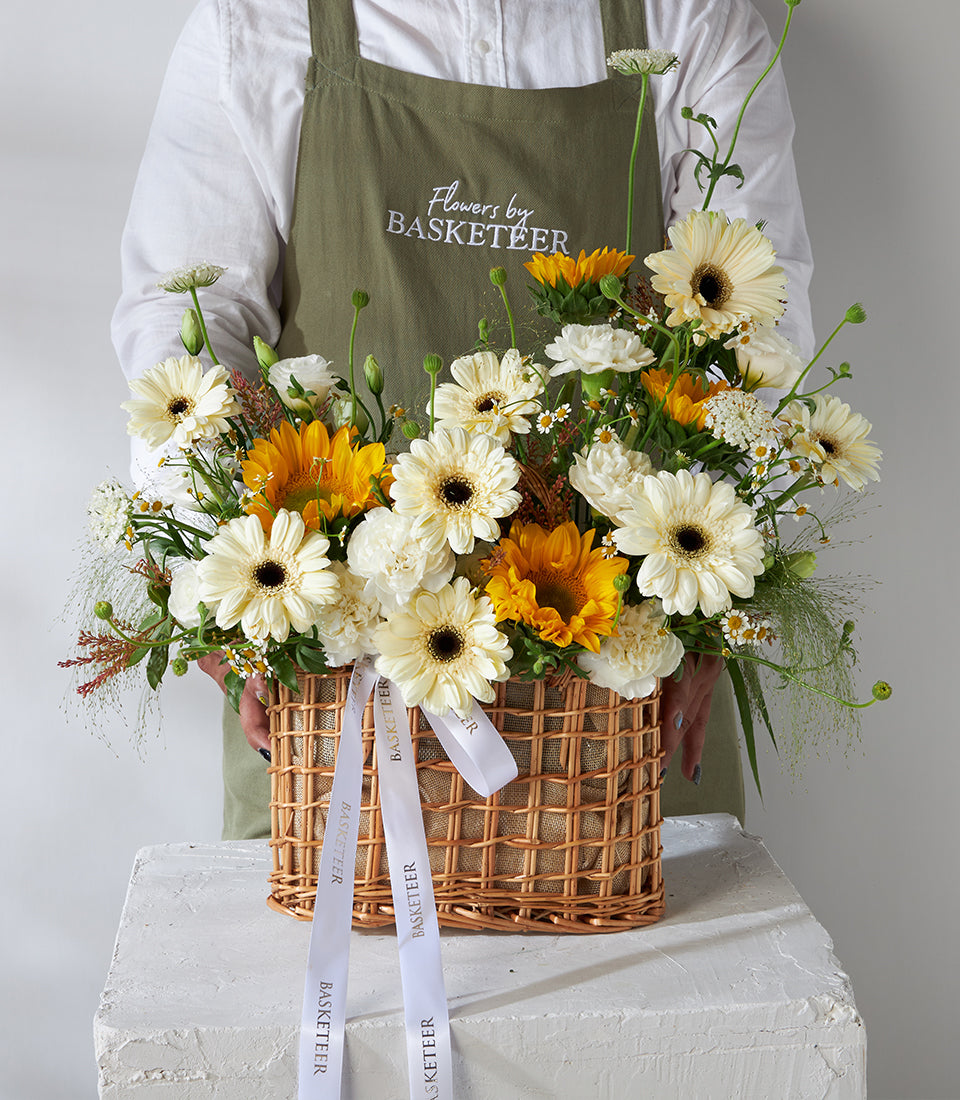 Florist holding wicker basket with yellow sunflowers, white gerberas, and carnations birthday gift arrangement