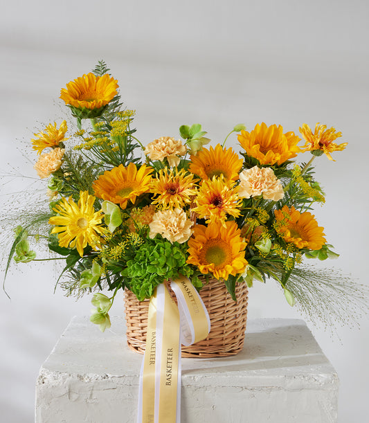 Vibrant yellow sunflowers and orange carnations in a woven wicker basket with cream ribbon