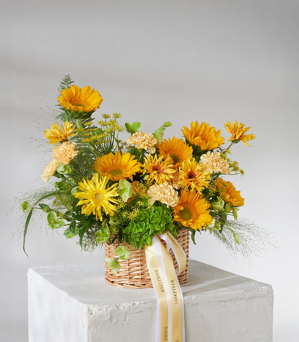 Yellow sunflowers, carnations and chrysanthemums in woven wicker basket with green foliage