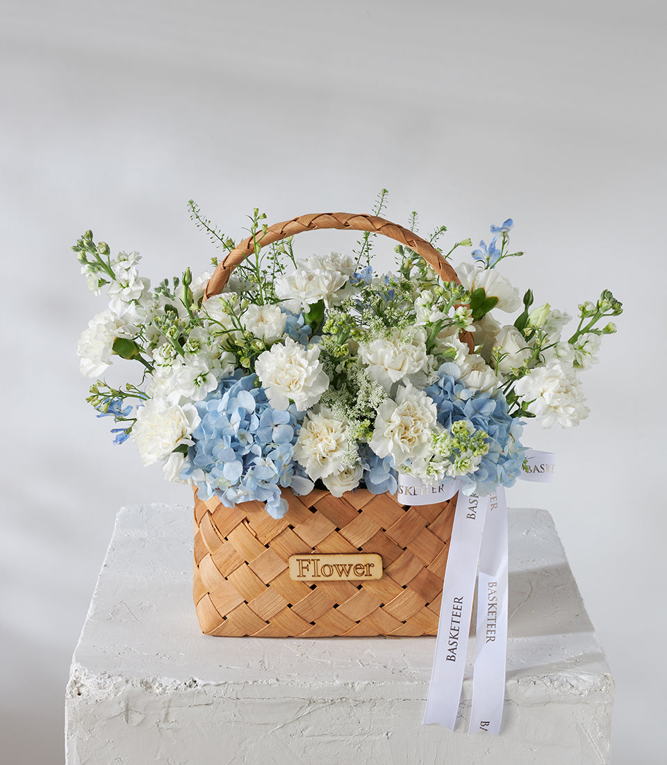 White carnations and blue hydrangeas in woven basket with satin ribbon, fresh birthday flowers arrangement