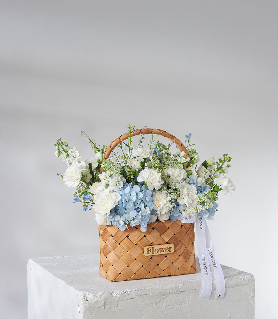 White carnation and blue hydrangea birthday bouquet in woven basket with green foliage
