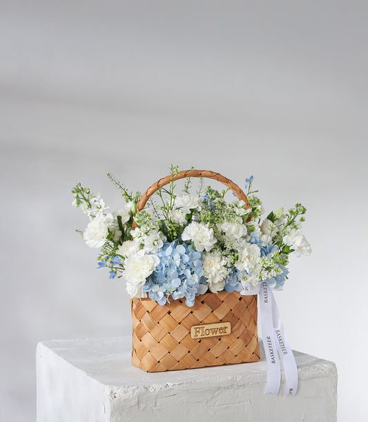 White carnation and blue hydrangea birthday bouquet in woven basket with green foliage