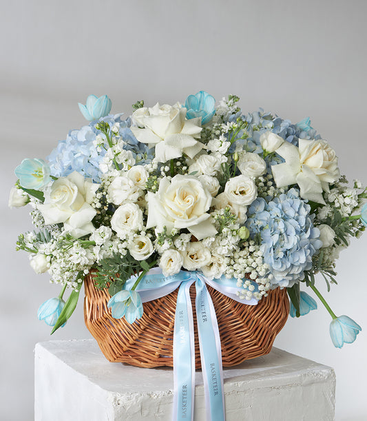 White roses, blue hydrangeas and tulips in woven wicker basket with fresh blooms and blue ribbon bow
