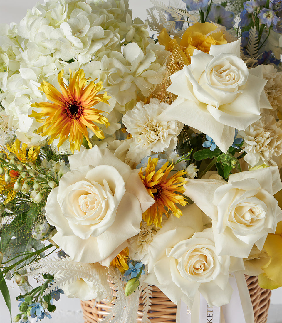 White and yellow roses with blue hydrangeas in woven basket arrangement