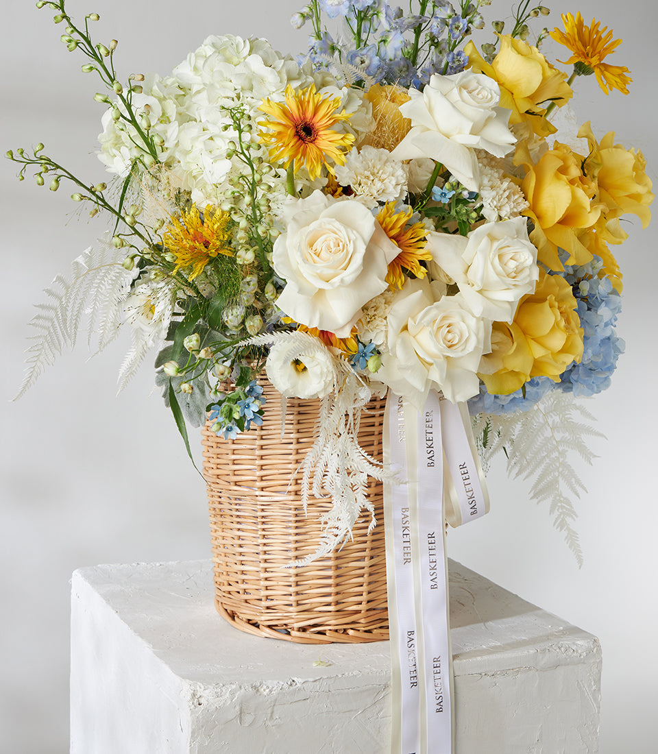 White and yellow roses with blue hydrangeas in woven wicker basket arrangement