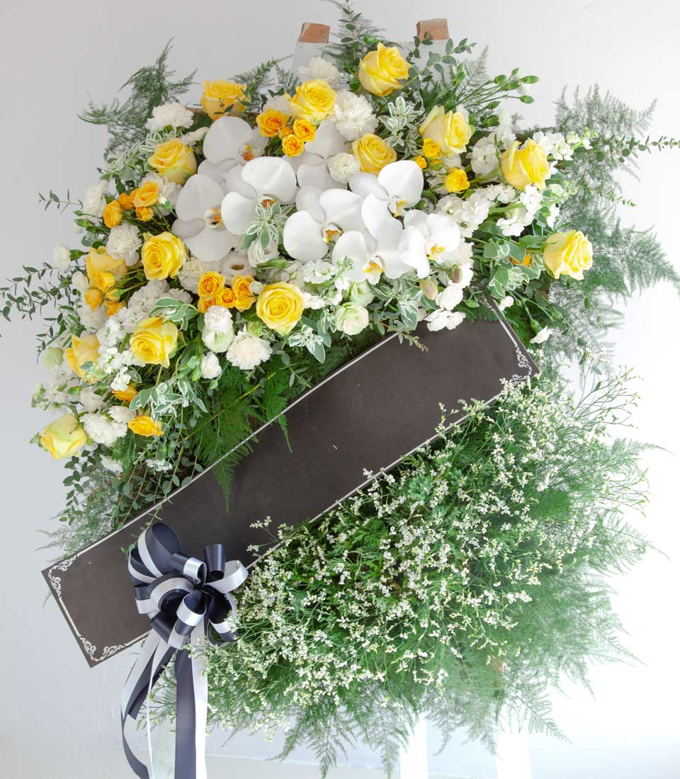 Tranquil Grace standing wreath with yellow roses, white orchids, and carnations on easel with tribute ribbon