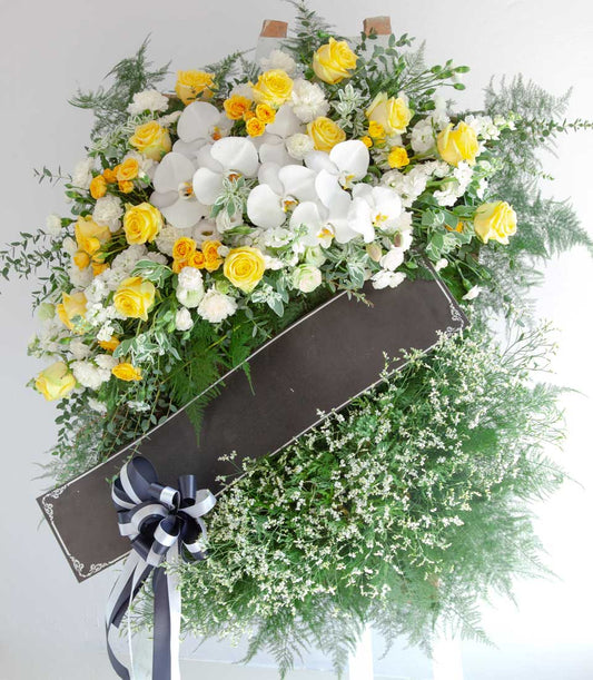 Tranquil Grace standing wreath with yellow roses, white orchids, and carnations on easel with tribute ribbon