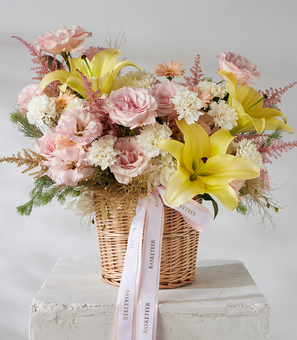 Yellow lilies and pink roses in woven basket with white ribbon bow, premium fresh flower gift arrangement