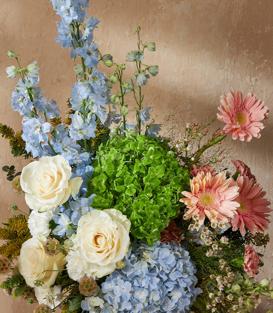 Lush garden bouquet with blue delphiniums, green hydrangeas, cream roses and pink gerberas in rounded vase arrangement