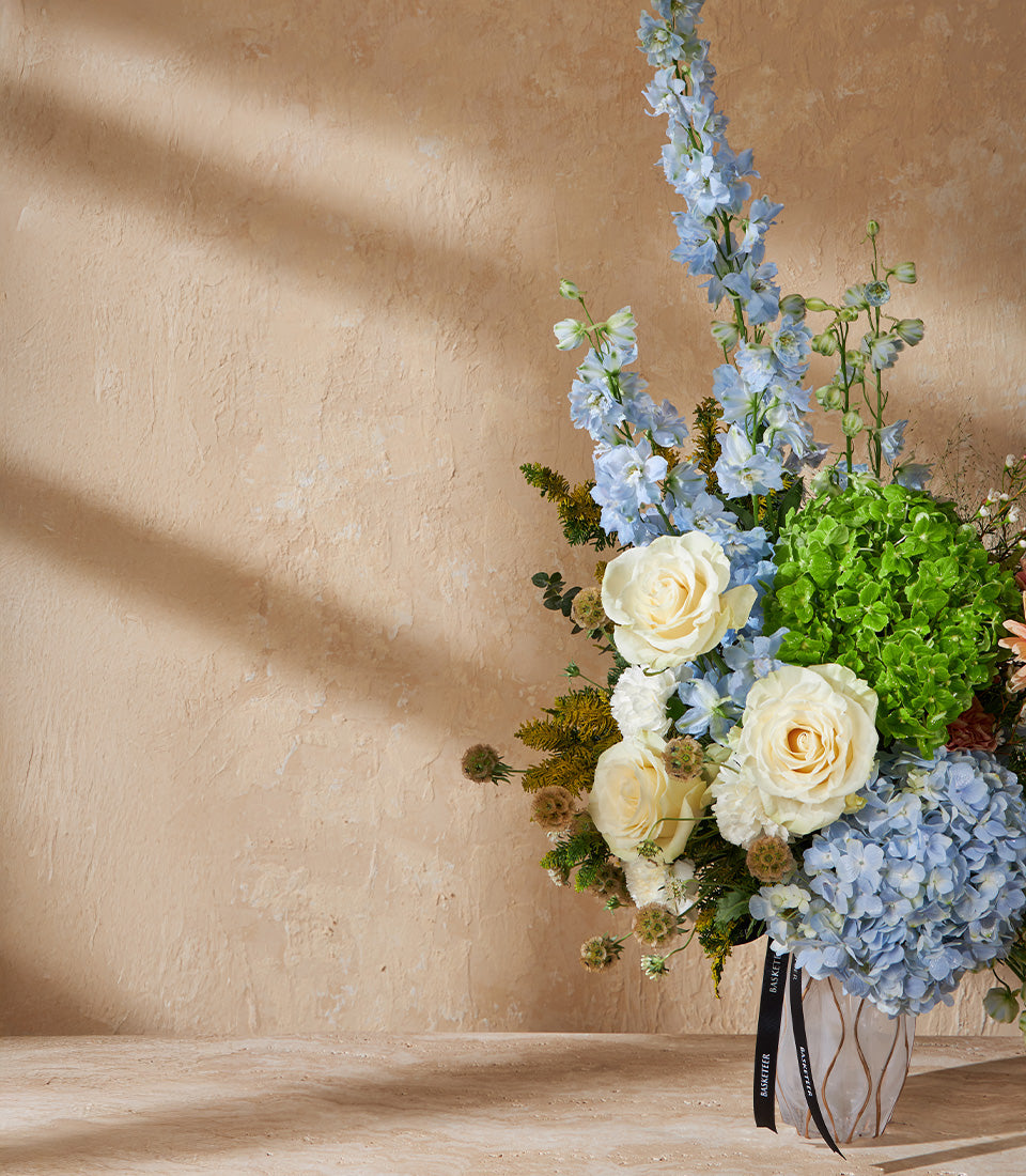 Light blue delphinium and hydrangea arrangement with creamy roses in textured ceramic vase
