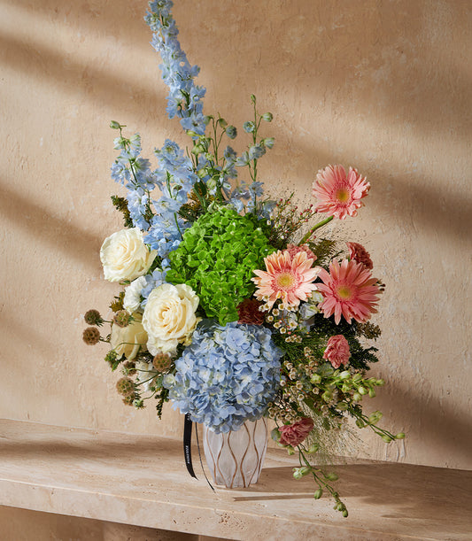Vibrant garden-style vase arrangement with blue delphiniums, white roses, green and blue hydrangeas, and pink gerbera