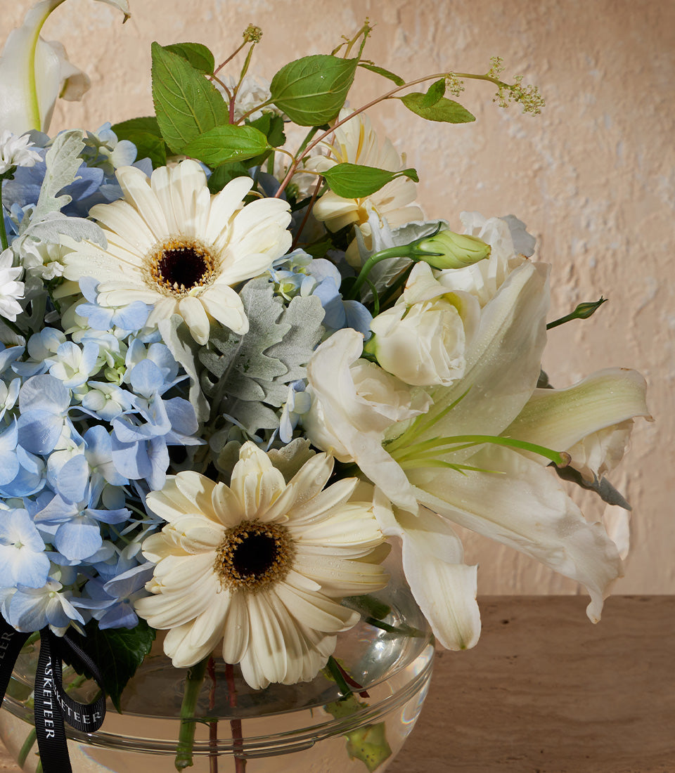 White and light blue gerbera daisies, hydrangeas, and roses in clear glass vase with green foliage