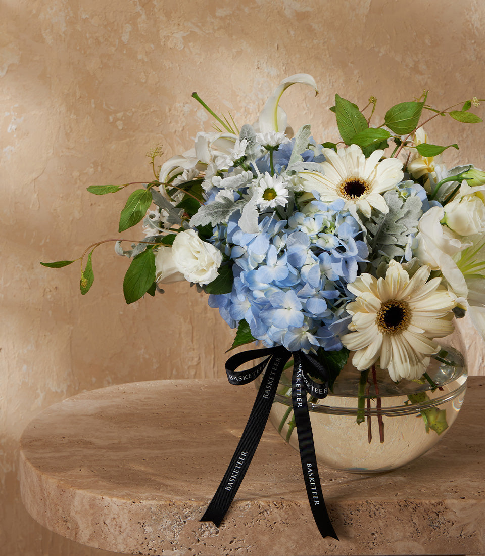 Blue hydrangea and white rose arrangement in spherical glass vase with cascading ribbon