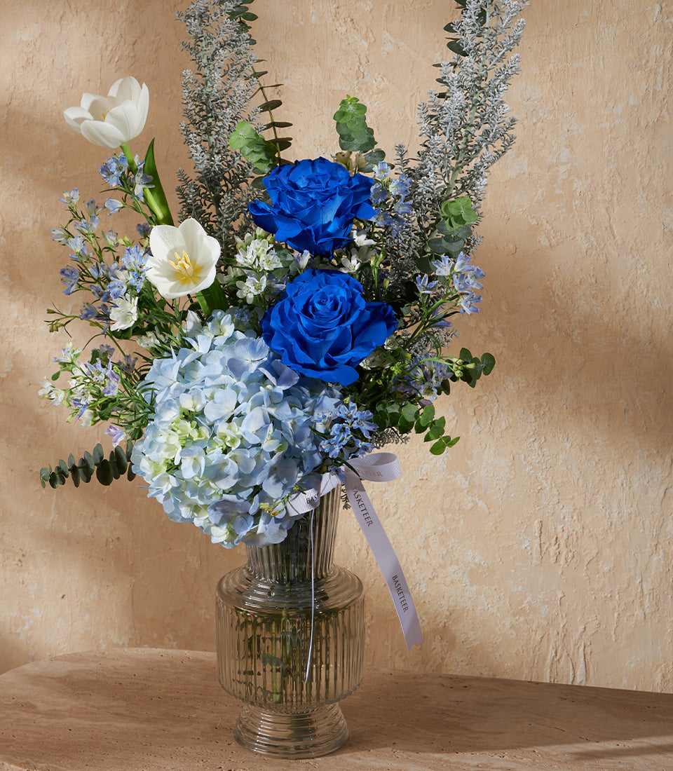 Deep blue roses and white tulips in ribbed glass vase with hydrangeas and eucalyptus greenery