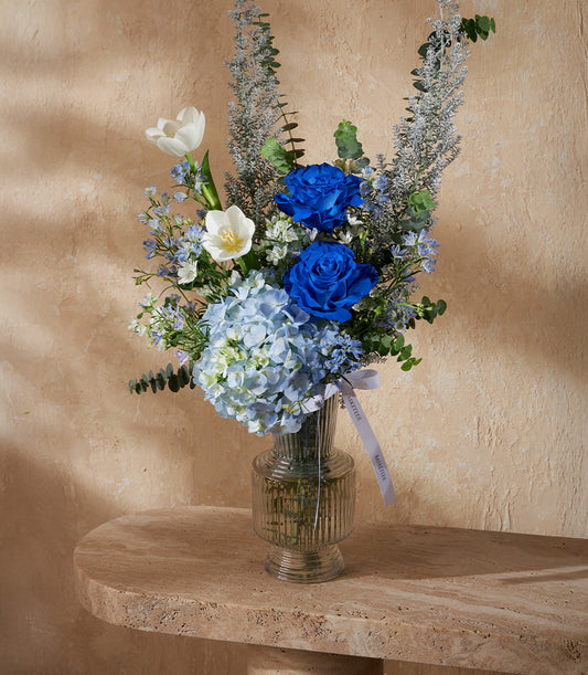 Blue and white rose and hydrangea arrangement in ribbed glass vase with white ribbon