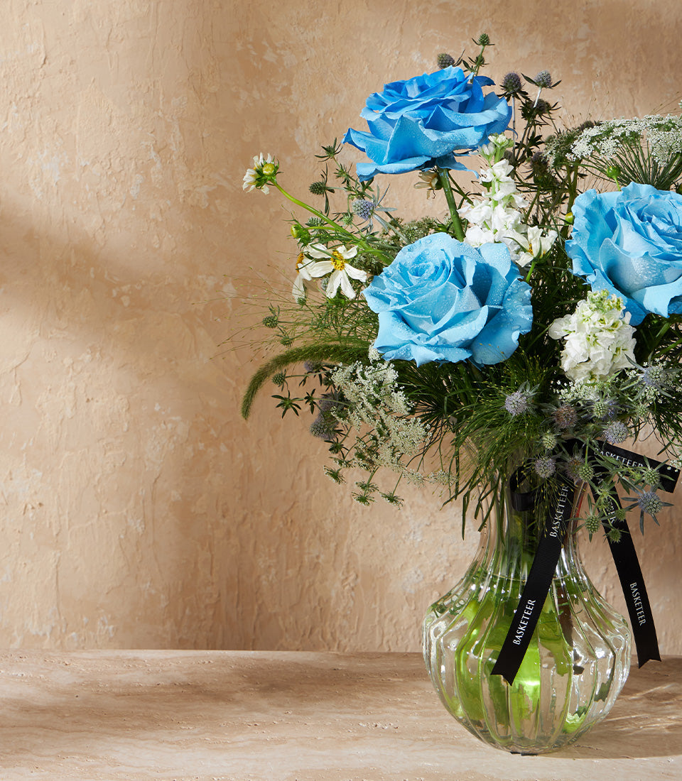 Fresh blue roses and white flowers in ribbed glass vase with green foliage and thistles