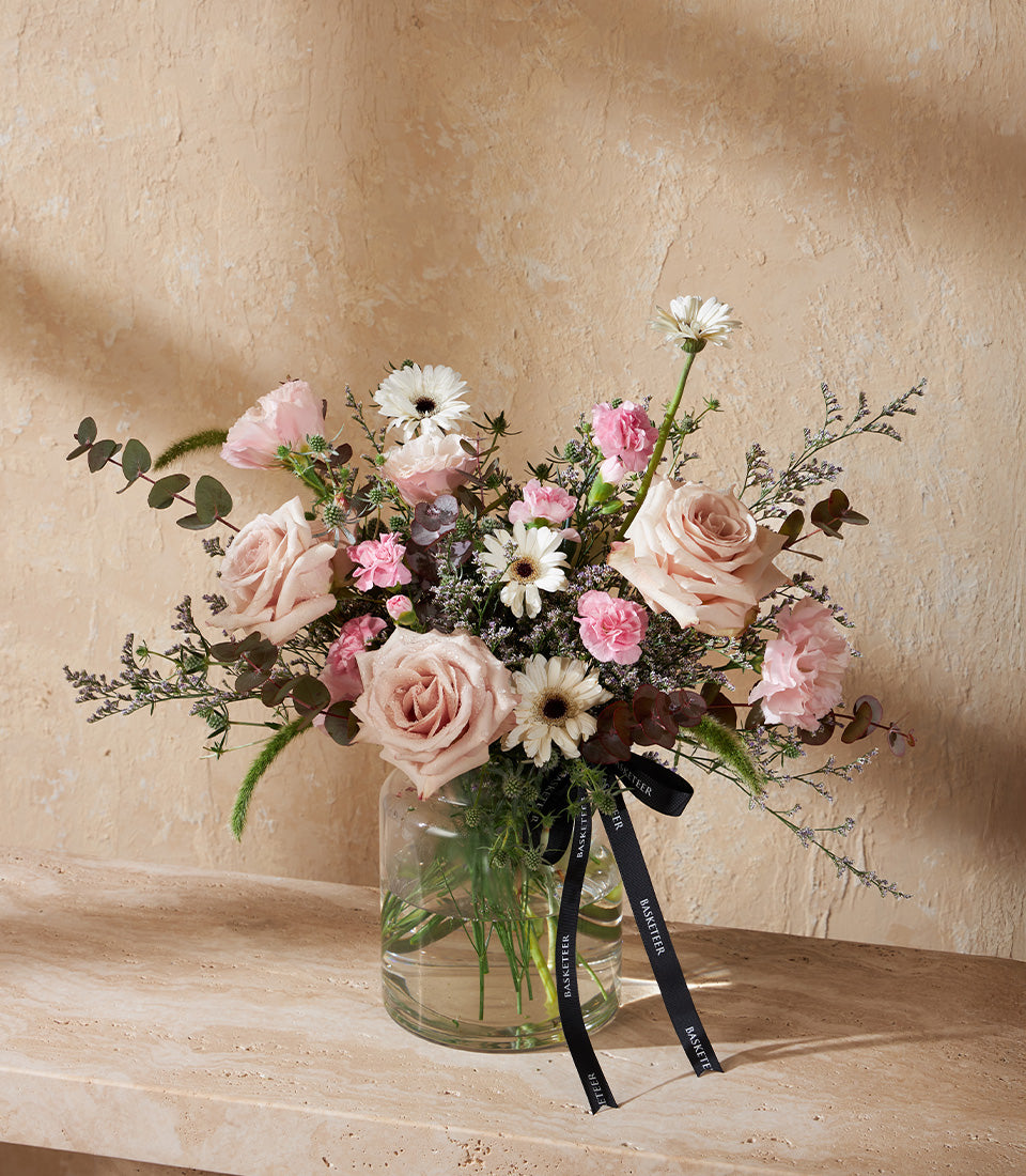 Light pink roses and yellow gerbera daisies in clear glass vase with green foliage and black ribbon