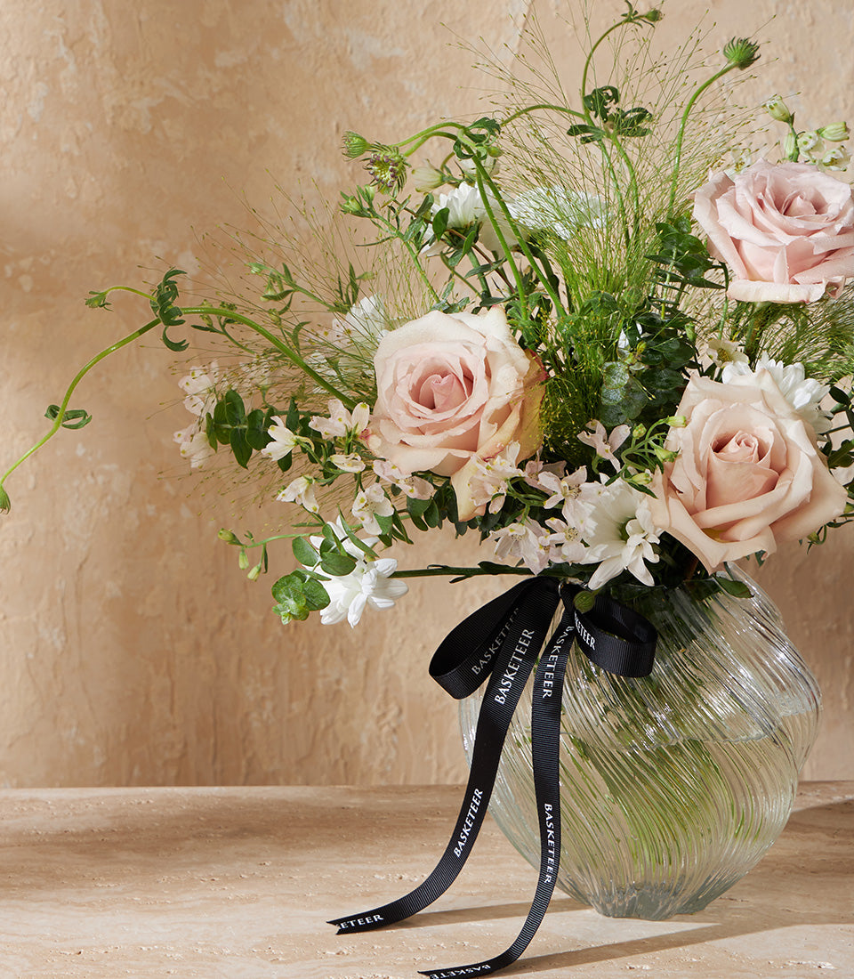 Blush pink roses and white flowers in textured glass vase with green foliage, black Basketeer ribbon