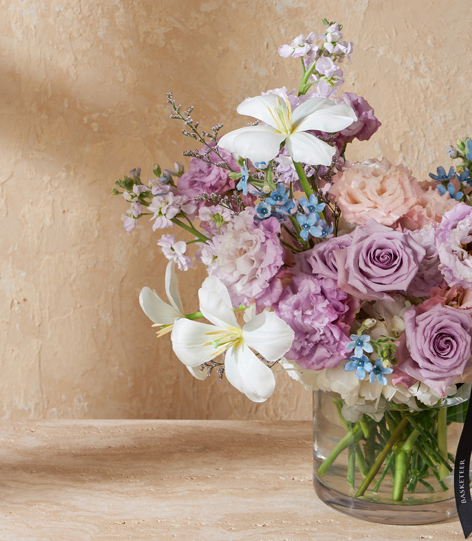 Ethereal lavender and white lily bouquet in glass vase with purple roses and delicate blue flowers
