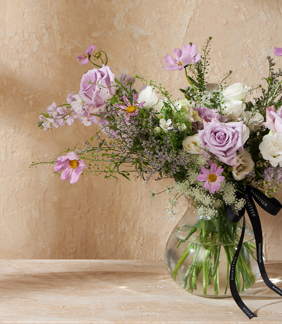 Lavender and white rose arrangement in clear glass vase with green stems and purple filler flowers