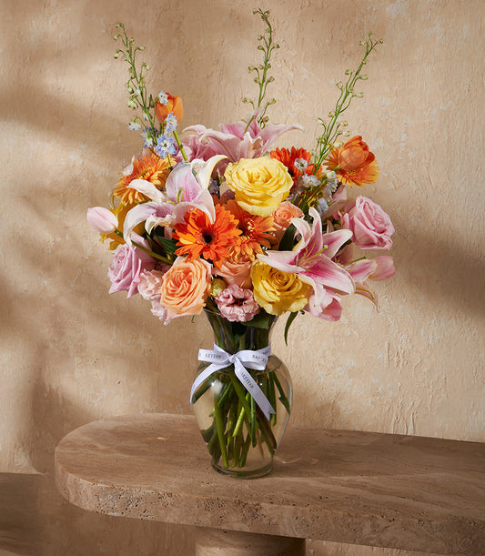 Radiant mixed flower arrangement with yellow roses, peach blooms, and orange gerberas in clear glass vase with white ribbon