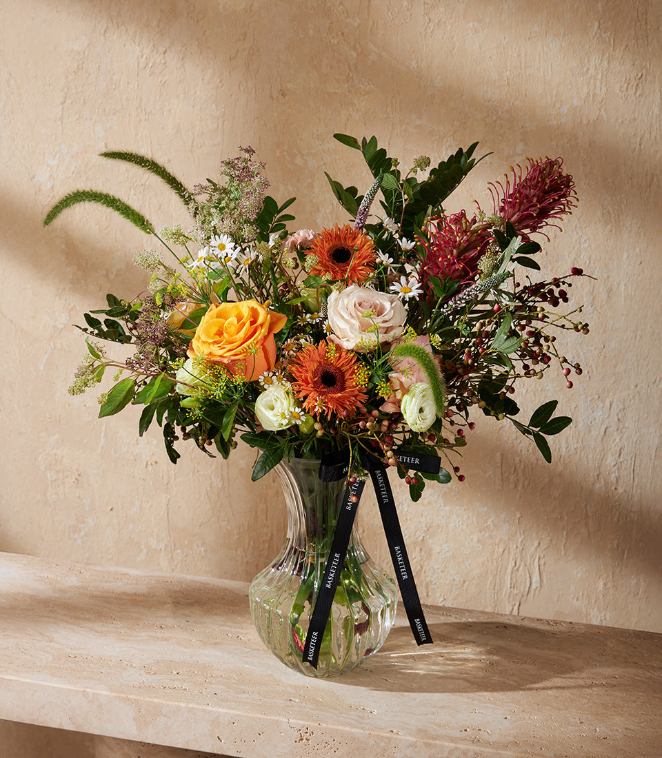 Vibrant mixed flower arrangement with orange roses, peach gerberas, and white ranunculus in ribbed glass vase