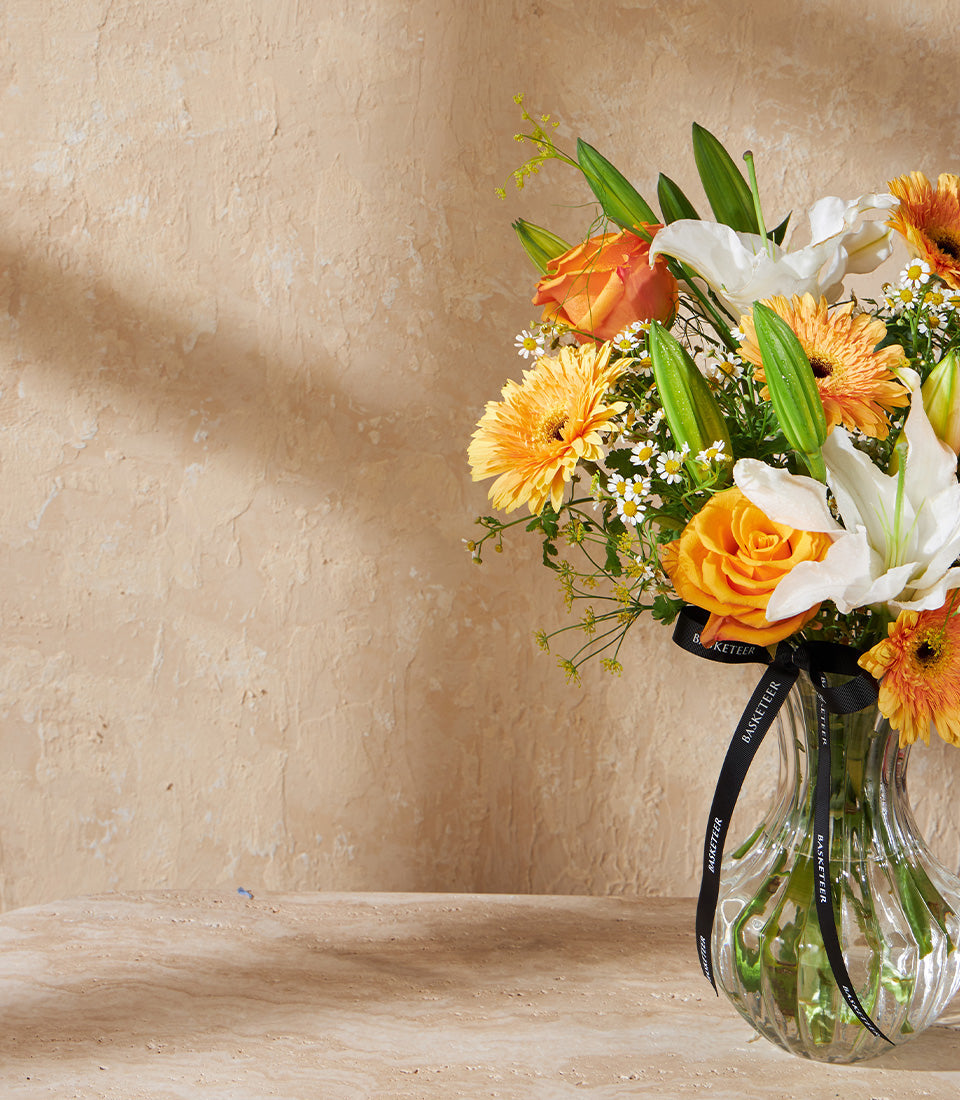 Sunlit Grace floral arrangement with orange and yellow roses, white lilies, and gerberas in clear glass vase