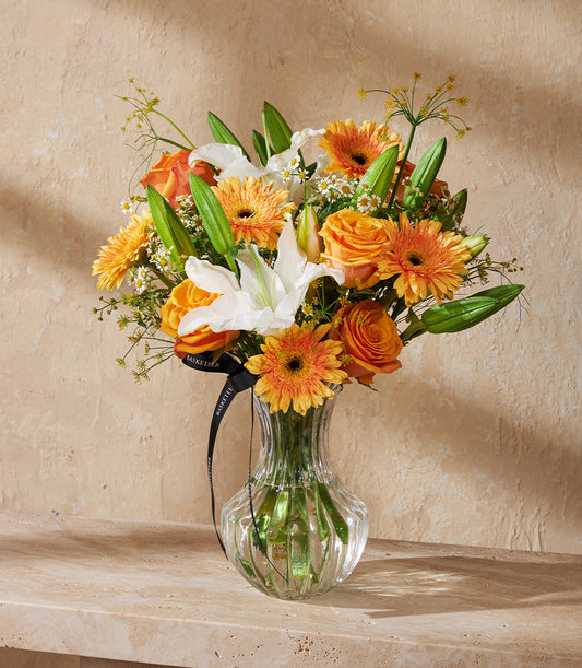 Vibrant orange roses, gerberas, and white lilies in a ribbed glass vase with fresh green stems