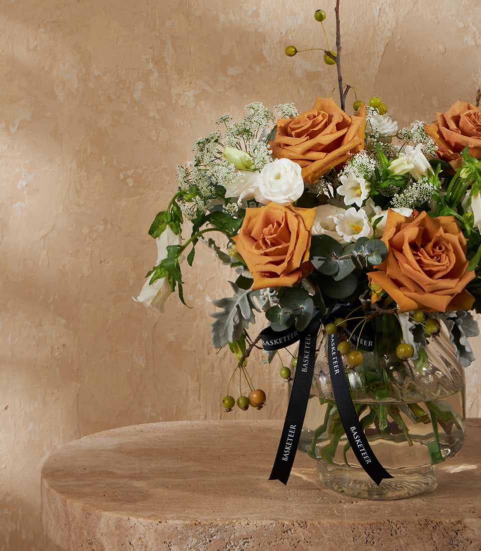 Burnt orange roses and white flowers in clear glass vase with eucalyptus, black Basketeer ribbon