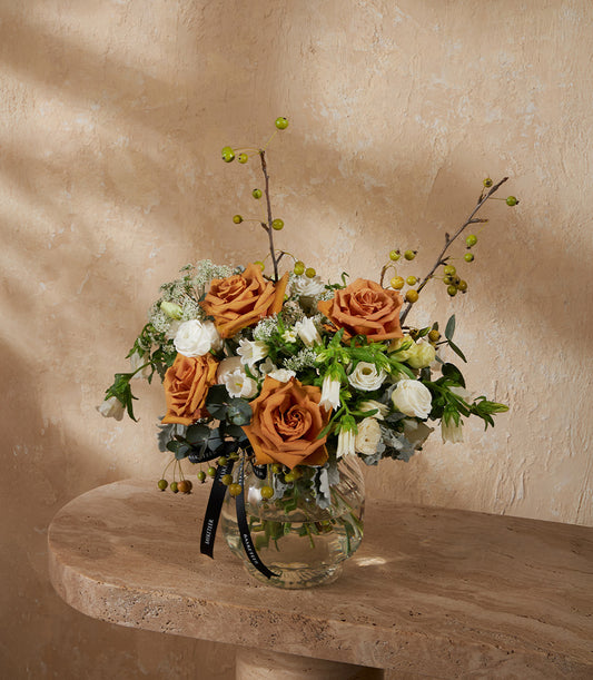 Rust-orange roses and white flowers in clear glass vase with green foliage and black ribbon