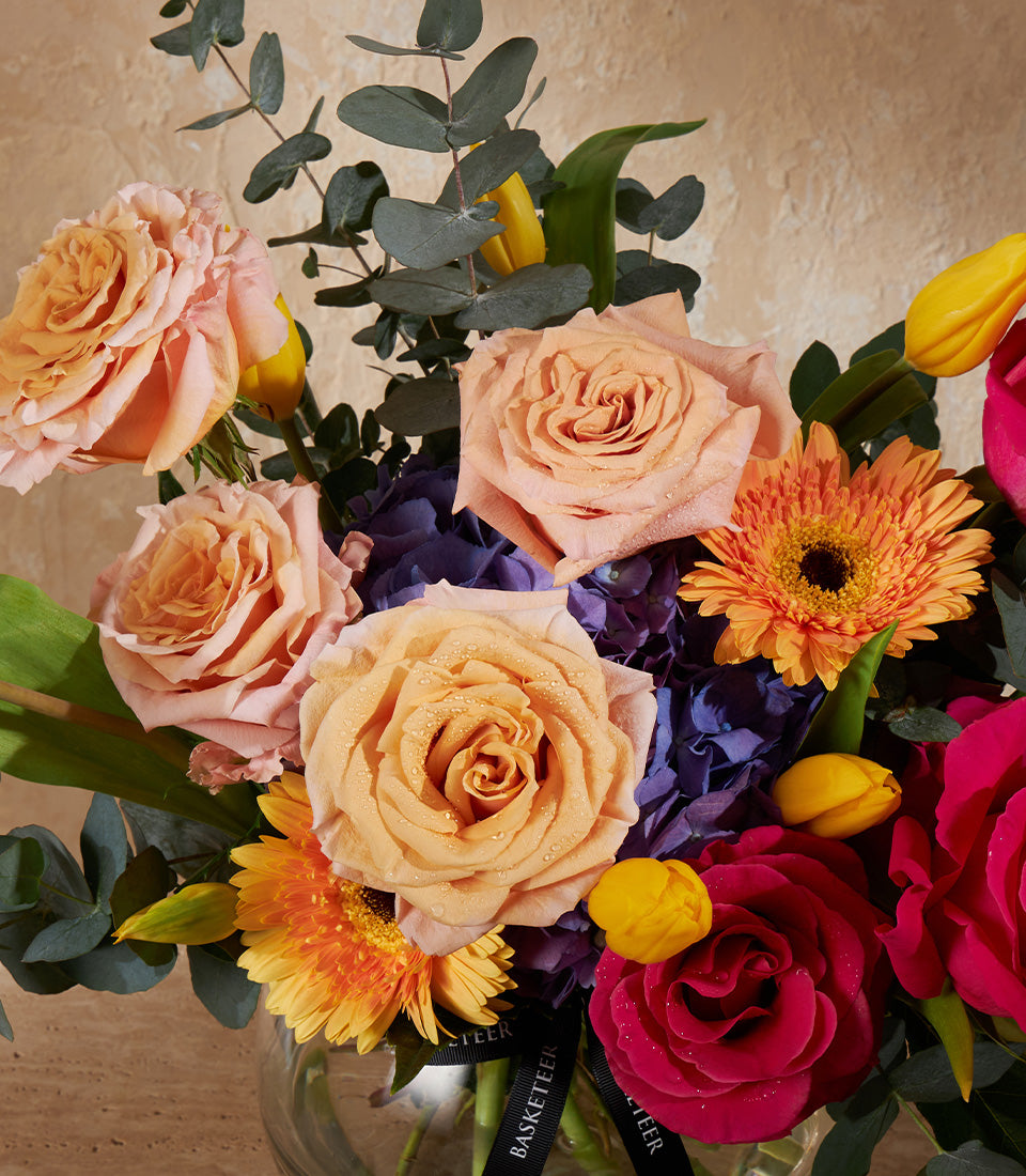 Radiant peach, pink, and red roses with orange gerberas and purple hydrangeas in clear glass vase