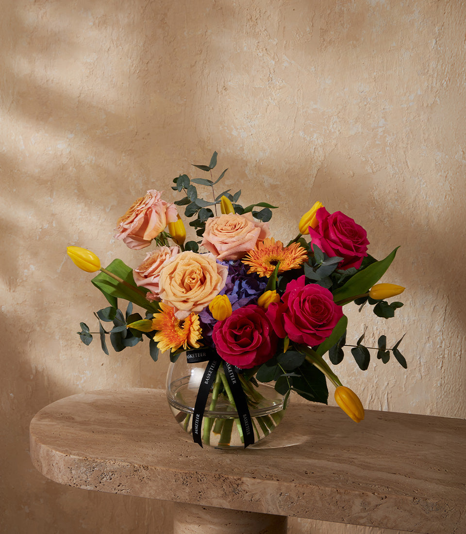 Radiant mixed flower vase with peach roses, yellow tulips, orange gerberas, and purple hydrangeas in clear glass sphere