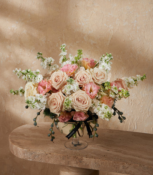 Light pink and cream roses with white filler flowers in a clear glass footed vase, tied with black ribbon