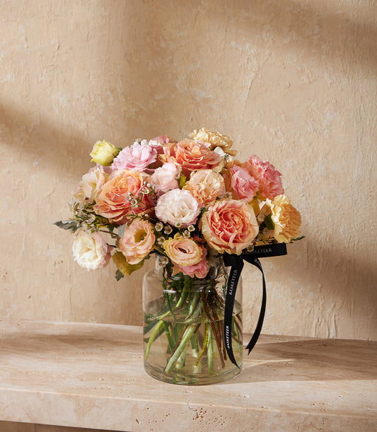 Peach and pink rose arrangement in clear glass vase with cream accents and premium ribbon