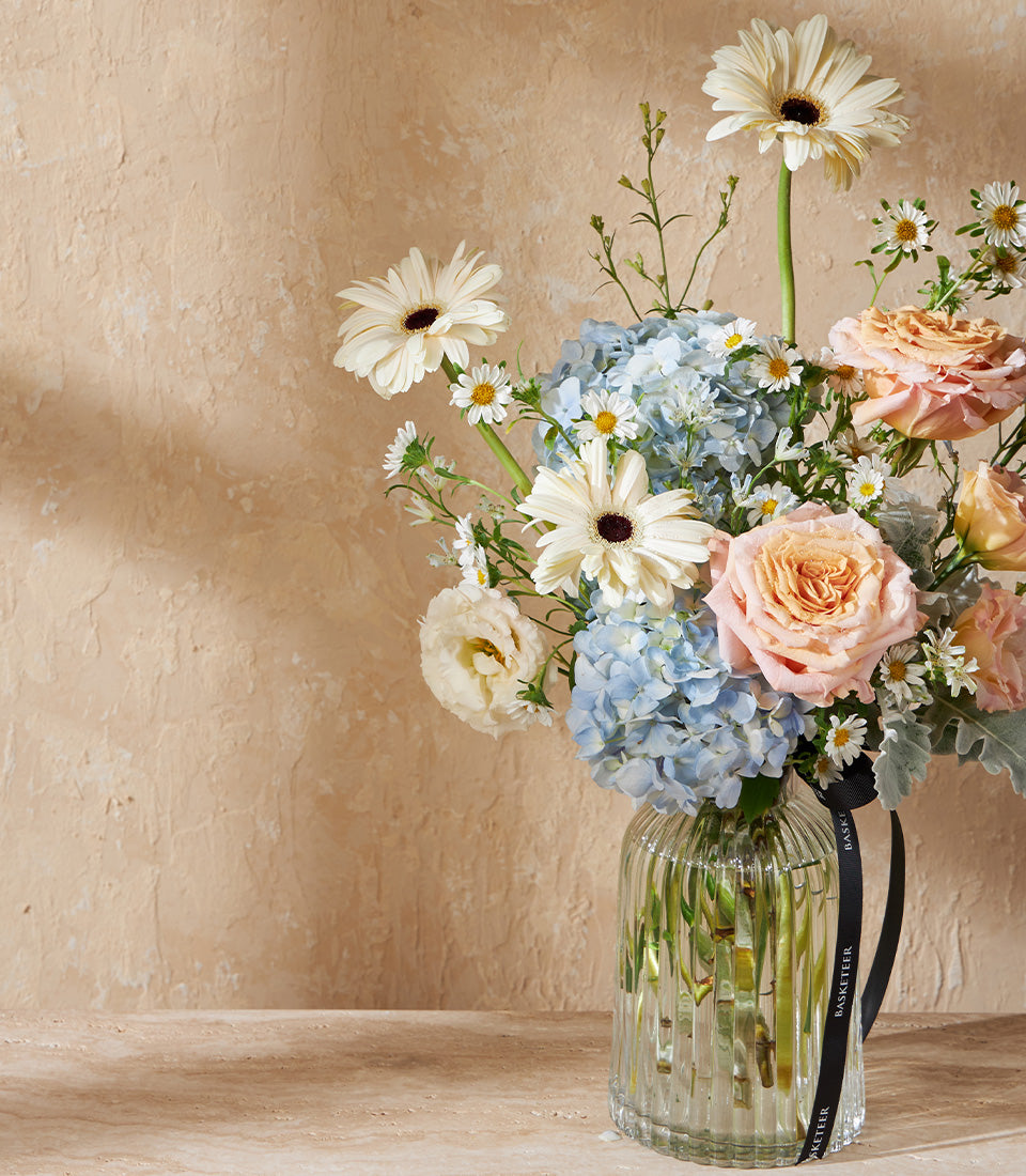 Vibrant mixed flower arrangement with white gerberas, blue hydrangeas, peach roses, and ranunculus in clear ribbed glass vase