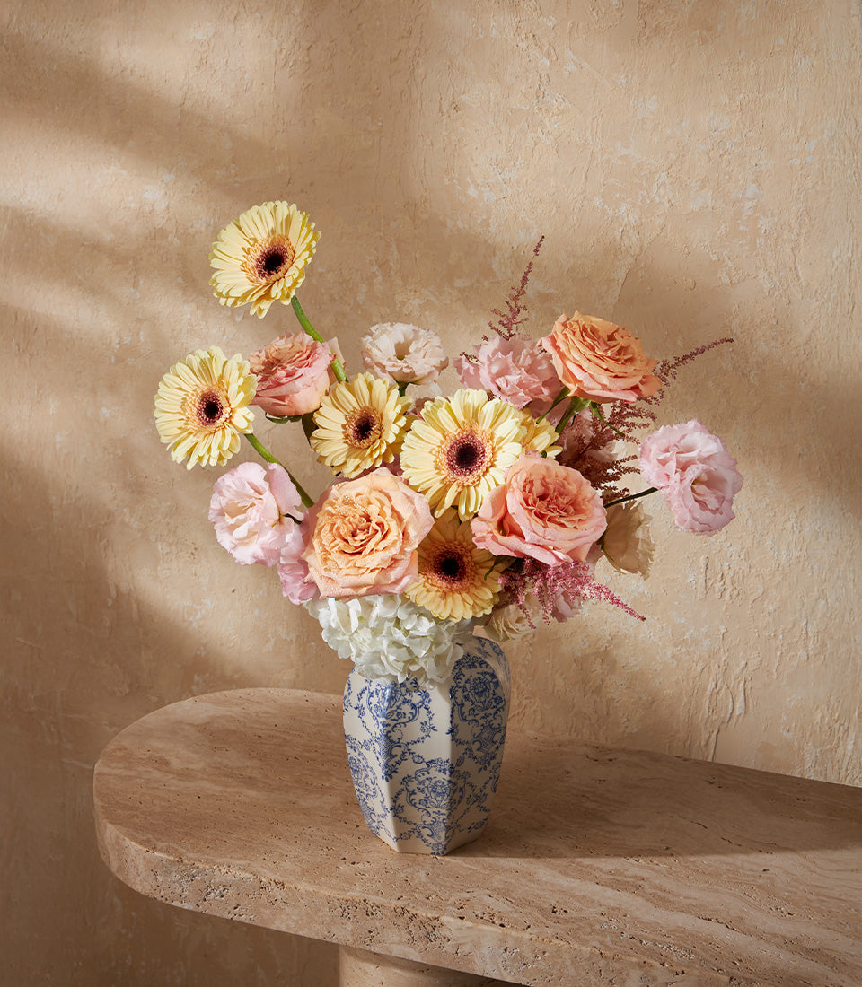 Yellow gerbera daisies, peach roses, and pink lisianthus in patterned white hexagonal vase
