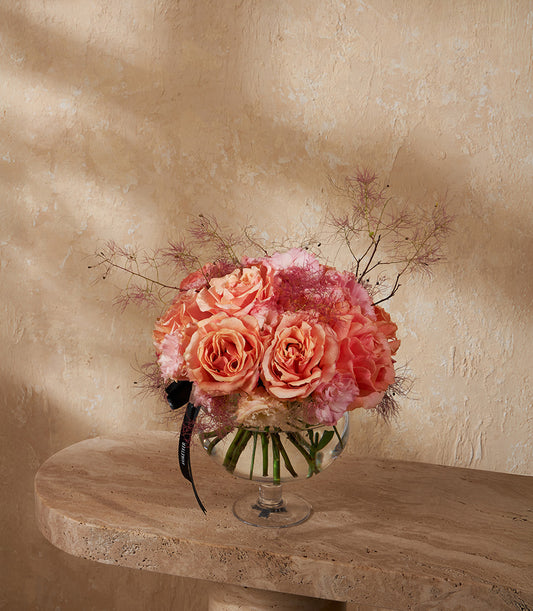 Peach and coral roses in clear glass vase with delicate foliage and ribbon accent
