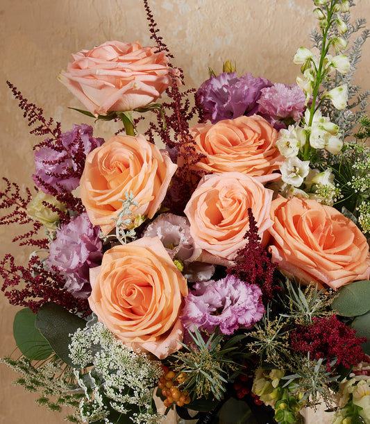 Peach and pink roses with purple lisianthus in a lush vase arrangement, featuring astilbe and eucalyptus